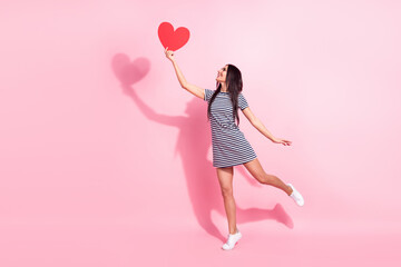 Full length body size view of pretty cheerful woman holding heart card having fun jumping isolated over pink pastel color background