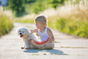 Cute blond child, toddler boy and pet dog, eating watermelon in garden