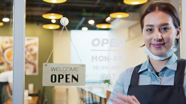 Portrait of Asian female restaurant owner wear mask, arms crossed and smile with open welcome sign at restaurant entrance door, slow motion. Business reopen after covid-19 coronavirus pandemic concept