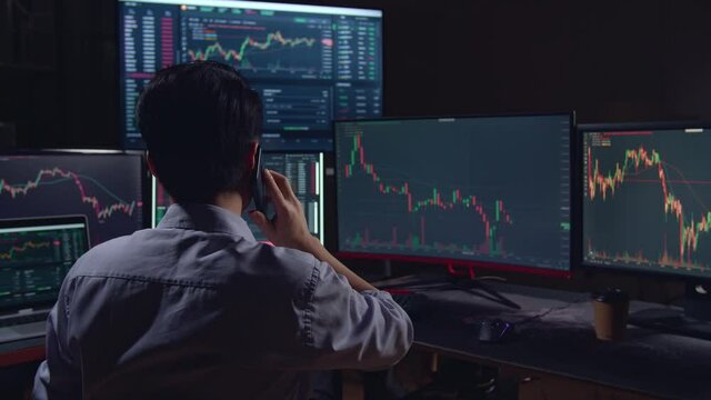 Back View Of A Asian Young Male Stock Market Broker Talking On Mobile Phone With Analyzing Graphs On Multiple Computer Screens
