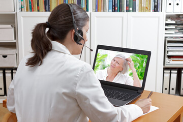 Doctor with headset and laptop, taking notes during a video call with a female patient suffering...