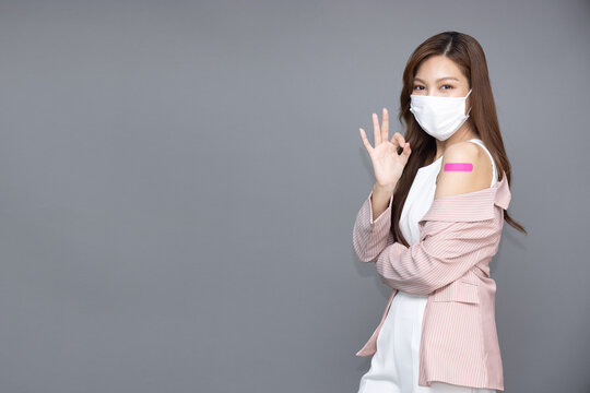 Beautiful Asian Woman Wearing Face Mask And Showing Ok Sign And Arm With Plaster Or Bandage Of Covid-19 Vaccinated Isolated On Grey Background