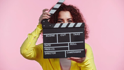 curly young woman obscuring face with clapboard isolated on pink