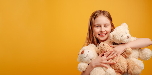 Beautiful girl child with three teddy bears in her hands isolated on yellow background with copyspace. Horizontal portrait of kid holding toys and smiling looking at the camera