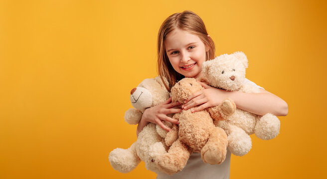 Cute girl child hugging three teddy bears isolated on yellow background with copyspace. Horizontal portrait of kid holding toys and smiling looking at the camera