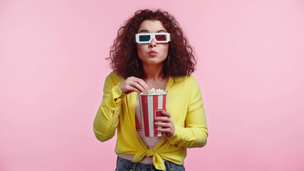 curly young woman in 3d glasses holding bucket while eating popcorn isolated on pink