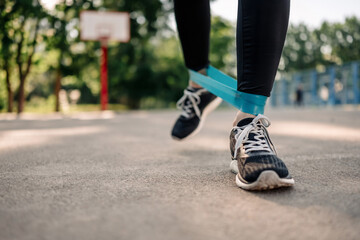 Fototapeta na wymiar Young girl woman exercising outdoors with rubber elastic band doing workout for legs. Closeup view of active training with additional sport equipment outside