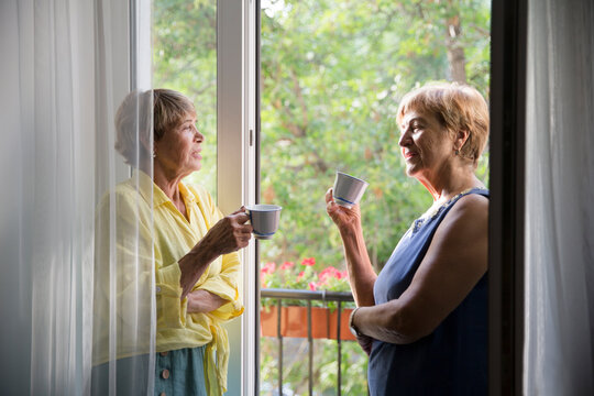 Portrait of happy senior woman friends standing on the balcony of the apartment and drink tea and talk

