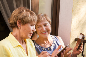 happy senior woman friends using smartphone, looking at screen and talk on the balcony of the apartment
