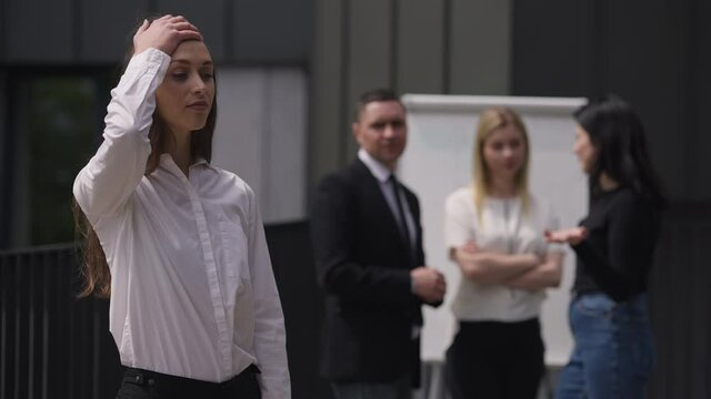 Arrogant burnt out young woman shaking head making facepalm gesture standing on business meeting with coworkers talking at background. Portrait of egoistic Caucasian millennial posing outdoors