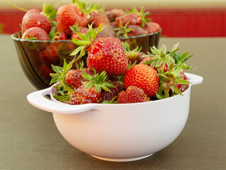 a lot of strawberries with green tails in a plastic bowl and a glass bowl behind. berry picking and harvest concept.