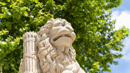 Obraz premium Light beige stone sculpture, representing a lion, close-up outdoors on a sunny day, green foliage and blue sky in the background