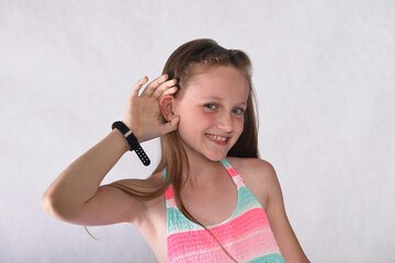 A beautiful school-age girl in the studio on a white background stands forward and holds her ear...