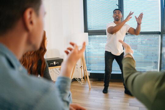 Portrait of cheerful African American male playing charades with friends showing pantomime by window in light office room. Diverse multi-ethnic colleagues playing in active games during team building.