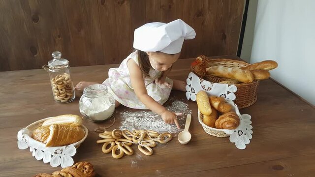 A three-year-old girl in a white dress and a baker's hat draws with her hands on flour scattered on the table, against the background of baskets with rolls, croissants, baguettes and bagels