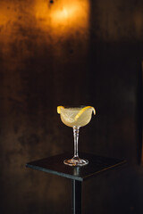 yellow cocktail champagne coupe lemon on pedestal against dark metal with orange glow still life 