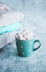 Turquoise cup, mug of hot drink with white marshmallow on the background of stack of winter knitted clothes. Winter cozy still life. Hot beverage - cacao, milk, cafe