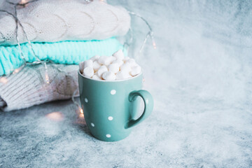 Obraz na płótnie Canvas Turquoise cup, mug of hot drink with white marshmallow on the background of stack of winter knitted clothes. Winter cozy still life. Hot beverage - cacao, milk, cafe