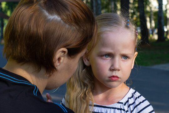 Close-up of frustrated little blonde preschool girl in a striped T-shirt with braided hair stands next to her mother and looks to the side in the park in good weather. The mother comforts her daughter