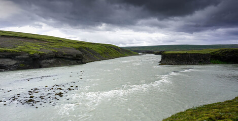 the river flows between green banks, cloudy rainy sky, nature of Iceland