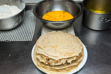 Ready-made pancakes are on the table, in a white plate, next to them are bowls with products for making pancakes: yolks, flour and butter.