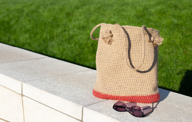 Brown knitted bag on concrete border in front of green grass in the park. Eco-friendly shopping jute bag. DIY. Zero waste lifestyle. - obrazy, fototapety, plakaty