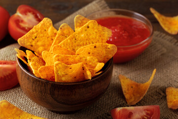 Corn chips at brown wooden bowl and tomato sauce in glass bowl on burlap cloth on brown table . Mexican tortilla meal with spices. Nachos with salsa