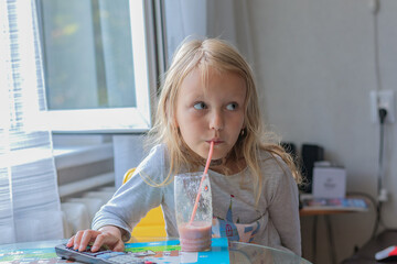 little girl with blond hair, 6 years old, sitting in the kitchen and drinking a milkshake with a straw from a glass glass, portrait of a child, pretty blonde preschool age, sitting at a table 