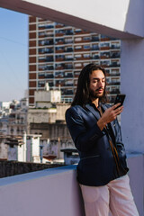 vertical photo of a young hispanic latin long haired business man talking using his smart phone, standing on a terrace overlooking the city.