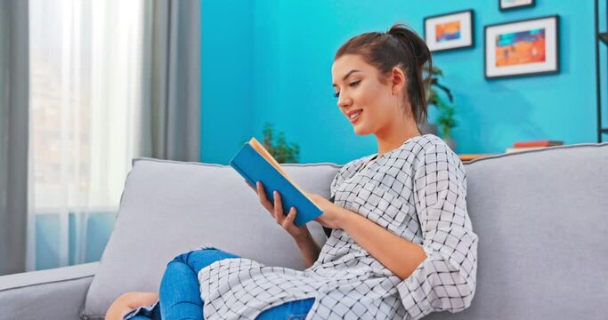Beautiful woman with hair pinned up relaxes in homely attire while delving into book that is in hands, reads page with curiosity, analyzing meaning of novel, turning pages without stopping reading