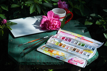 white sheet of watercolor album, lying on the table, surrounded by a cup of tea, watercolor set and bushes of peonies 