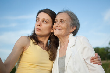 Senior mother with gray hair with her adult daughter looking at the camera in the garden and hugging each other during sunny day outdoors, mothers day
