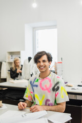 joyful fashion designer looking at camera near colleague working on blurred background