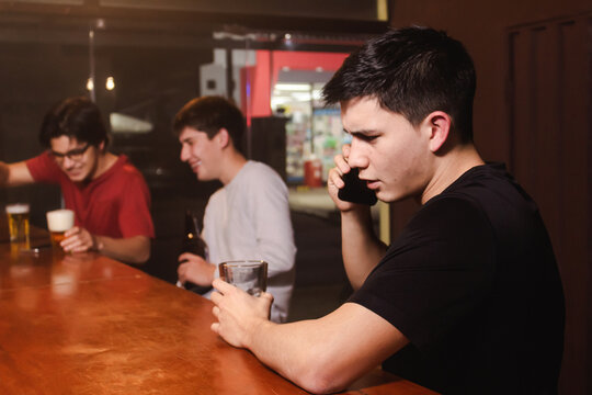 An angry young man talking on the phone while his friends enjoy a beer and laugh at the bar.