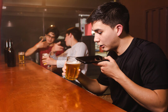 A young man sending a voice audio while drinking beer with his drunken friends at the bar.