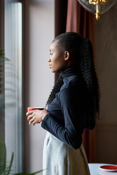 Fashionable African girl sitting at cafe table, enjoying morning cappuccino. Young black woman drinking coffee, contemplating urban views through window