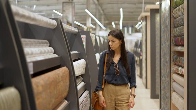 Young female customer looking at large assortment of wallpapers for home interior design in construction department of shopping mall, woman choosing materials for repairing her room