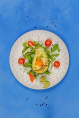 Fried eggs baked in half an avocado, served with mixed salad, cherry tomatoes and parmesan. Top view on a blue background, with space, vertical