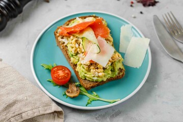Toast with scrambled eggs, avocado, smoked salmon, parmesan cheese and vegetables on rye bread. On a blue plate, horizontal