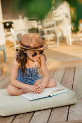 Mädchen mit Sonnenbrille sitzt auf Polster auf Holzterasse und liest ein Buch