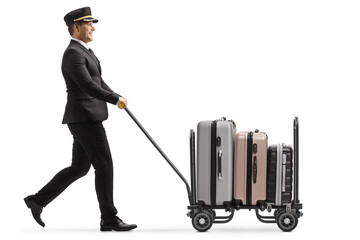 Full length profile shot of a bellboy walking and pushing a trolley cart with suitcases - obrazy, fototapety, plakaty