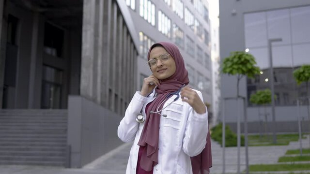 Beautiful young muslim female doctor wearing hijab and eyeglasses, smiling while posing at camera in her medical uniform and stethoscope in front of modern hospital building outdoors