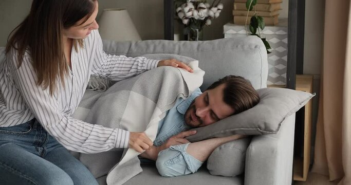 Caring young woman covering with plaid falling asleep husband on comfortable sofa in living room. Tired millennial guy daydreaming on soft pillow, enjoying lazy carefree relaxed weekend time.