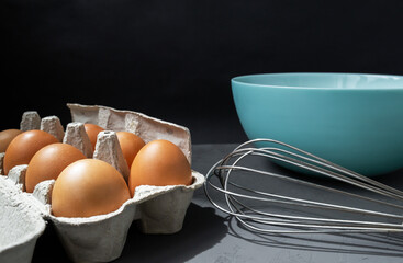 Fresh eggs in cardboard egg tray with whisk and plate on dark background