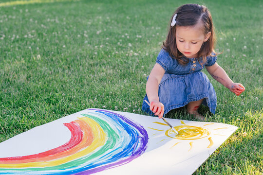 Little girl 2-4 years old paints rainbow and sun on large sheet of paper, sitting on green lawn