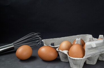 Fresh eggs in a cardboard egg tray with a whisk on a dark background