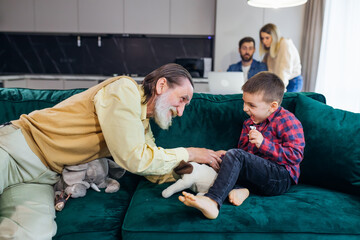 Old grandfather and little boy's grandson are playing toy game, sitting on the couch, funny little grandson having fun with his grandfather, in the background a couple is working on a laptop