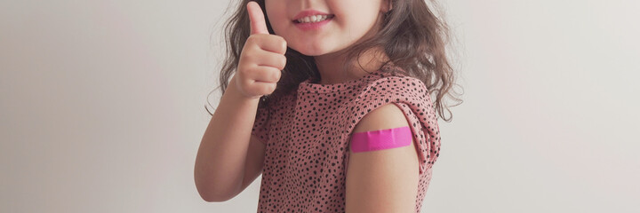 Young  girl giving thumb up and showing her arm with pink bandage after got vaccinated or...