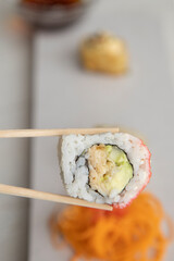 closeup to chopsticks holding a sushi roll with rice and seafood, detail of the texture in the background of a plate with more food, lifestyle