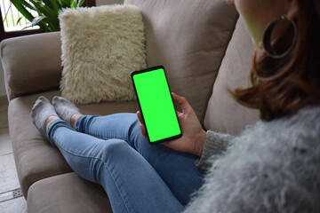 Image of a woman holding a mobile phone with a green screen while sitting on the sofa with a feeling of relaxation.